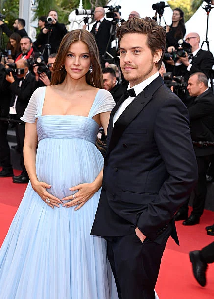 Close-up of Barbara Palvin in a pale blue pleated gown cradling her baby bump beside a man in a black tuxedo on a red carpet